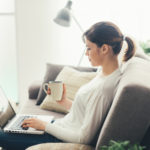Young woman relaxing at home on the couch, she is having a coffee and using a laptop