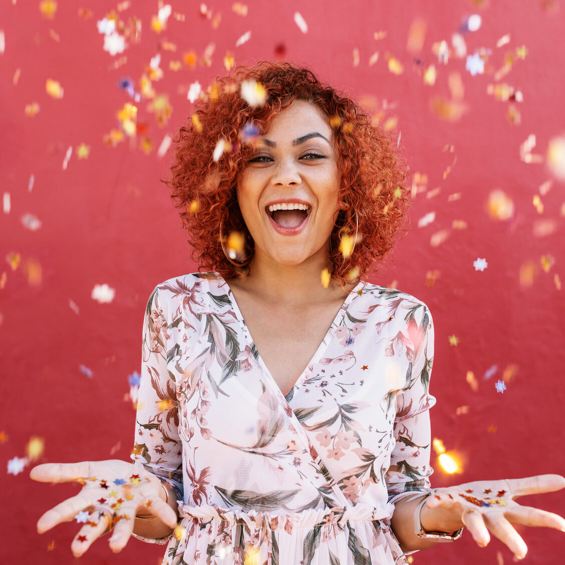 Young woman throwing star shaped colorful confetti in air. Close up of woman in happy mood with confetti all around against a red background.