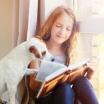 Child with dog reading book at home. Girl with pet sitting at window at read
